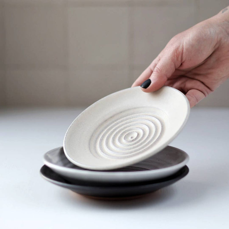 Gravesco Pottery - Garlic Grater Plate in Black. A hand lifts an off-white grater plate with concentric circles, ideal for preparing garlic and serving dips.