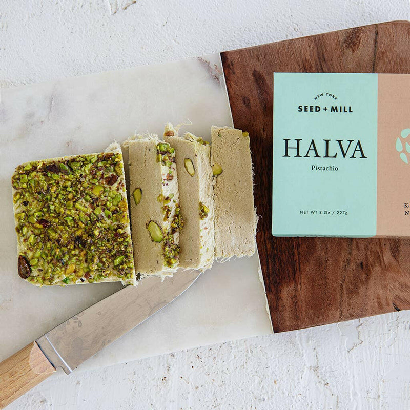 Seed + Mill | Halva - Pistachio: Close-up of flaky, sesame confection next to a wooden-handled knife, highlighting its texture. Ideal for desserts or charcuterie boards.
