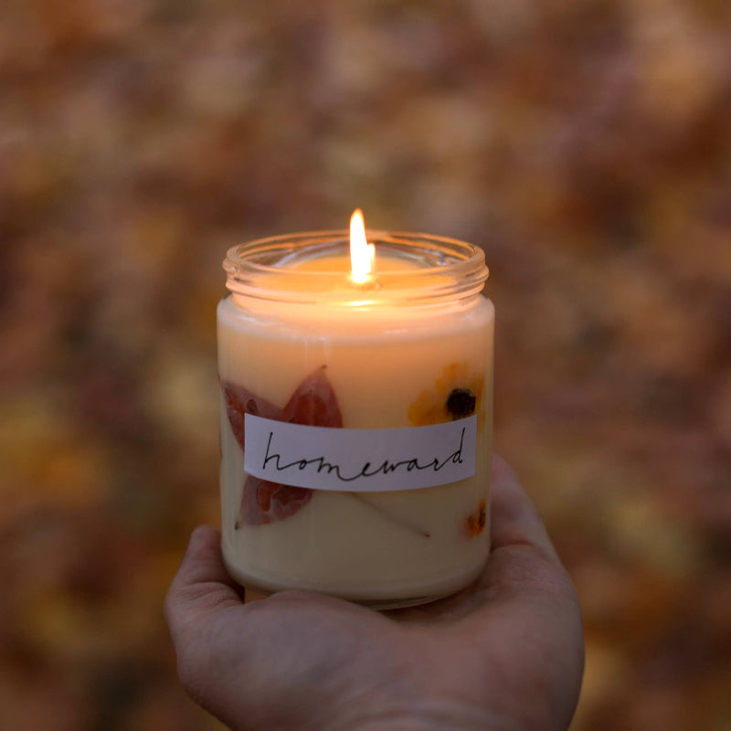 Hand holding Among the Flowers - Essential oil candle | HOLIDAY: Hearth in a glass jar, featuring pressed autumn leaves and blossoms, lit against a fall foliage background.