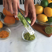Person zesting lime over bowl with Microplane | Classic Series Stainless Steel Zester, surrounded by citrus fruits, showcasing its zesting precision and kitchen utility.