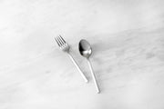 Zwiesel Fortessa | Arezzo Flatware Serving Set, Brushed: A stainless-steel fork and spoon with a geometric design on a marble-like surface, showcasing elegant craftsmanship.