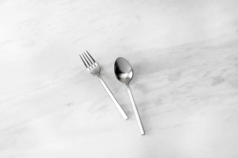 Zwiesel Fortessa | Arezzo Flatware Serving Set, Brushed: A stainless-steel fork and spoon with a geometric design on a marble-like surface, showcasing elegant craftsmanship.
