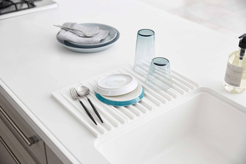 Yamazaki Home - Tower Folding Dish Drainer Mat in use, displaying plates, utensils, and glasses drying over a white kitchen sink countertop.