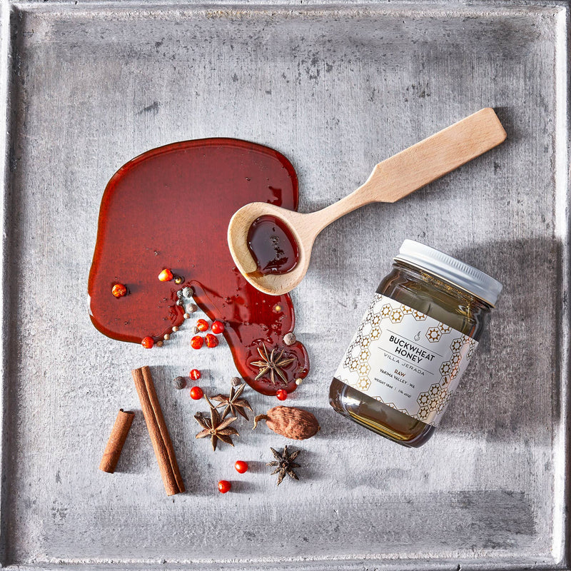 Villa Jerada - Buckwheat Honey - 1lb jar, tipped on a baking sheet with a wooden spoon and spilled honey, surrounded by spices like cinnamon, star anise, and peppercorns.