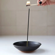 Hand ignites incense in Gravesco Pottery - Incense Burner, a black handmade dish, ideal for catching ash, enhancing room ambiance.