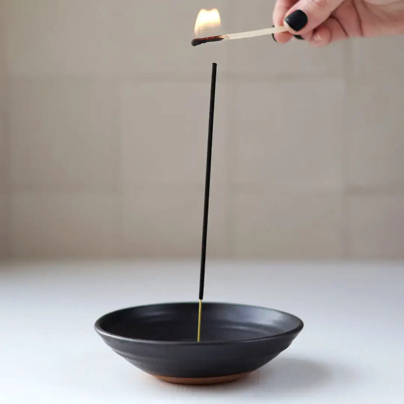 Hand ignites incense in Gravesco Pottery - Incense Burner, a black handmade dish, ideal for catching ash, enhancing room ambiance.