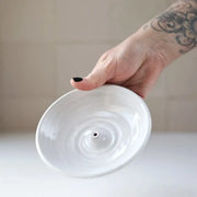 Hand holding Gravesco Pottery Incense Burner, a round, white-glazed ceramic dish designed for incense sticks, showcasing a central raised bump with a small hole.
