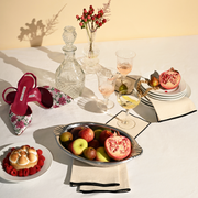 A vibrant tabletop features Manolo Blahnik shoes, fruit, drinks, and an elegant display of ATELIER SAUCIER - Cream Burlap Noir Cocktail Napkins in a culinary setting.
