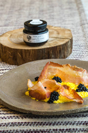 TartufLanghe Truffle Perlage pearls enhance creamy scrambled eggs with black caviar and crispy cured pork, showcased on a rustic plate alongside the product jar.