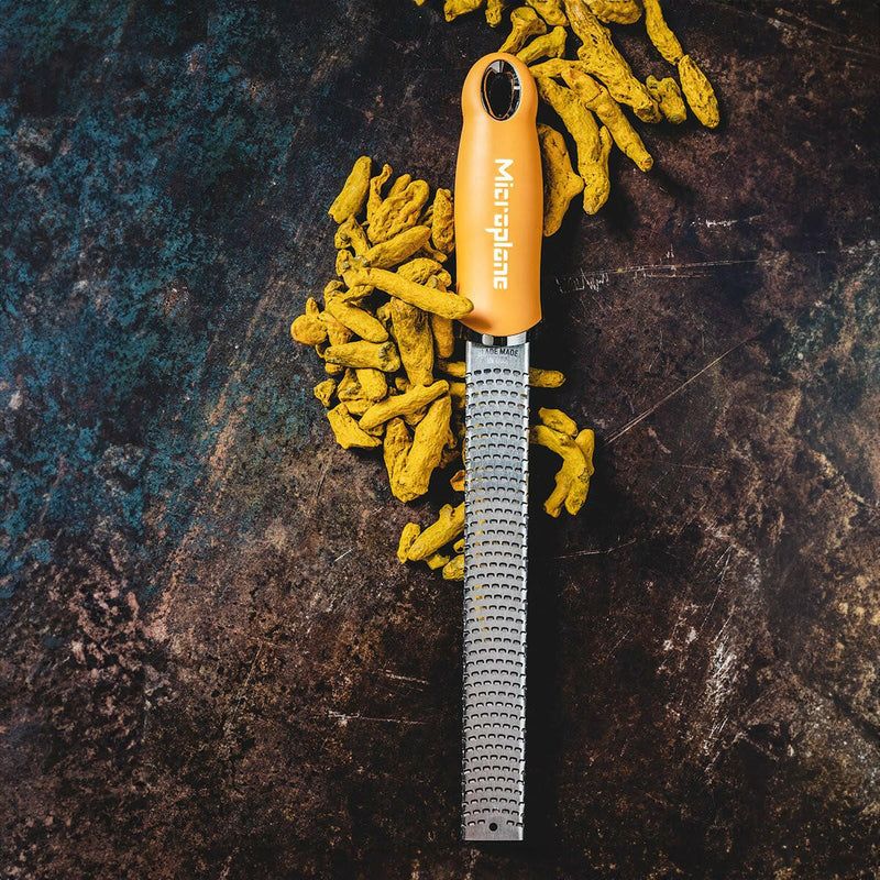 Microplane Premium Classic Zester Grater with orange handle, displayed with turmeric roots ready for grating, showcasing its effortless slicing capability.