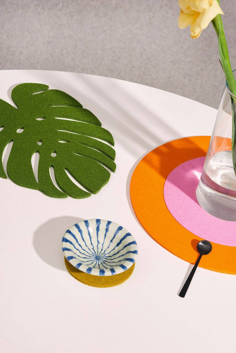 Graf Lantz - Merino Wool Felt Medium Monstera Leaf Trivet: Loden Green on table, alongside a vase and a glass, highlighting kitchen elegance.