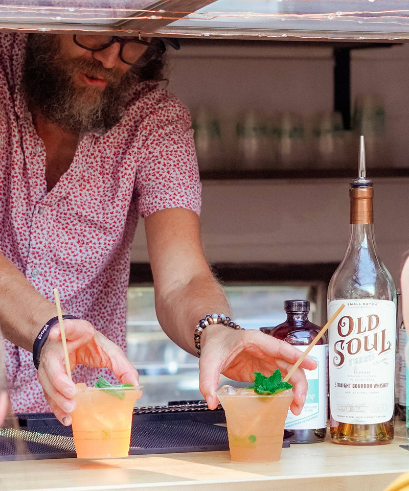 Bartender serves cocktails with mint garnish using Farmers Jam Peach Basil Cocktail Syrup, paired with Old Soul Bourbon, emphasizing easy, flavorful home mixology.