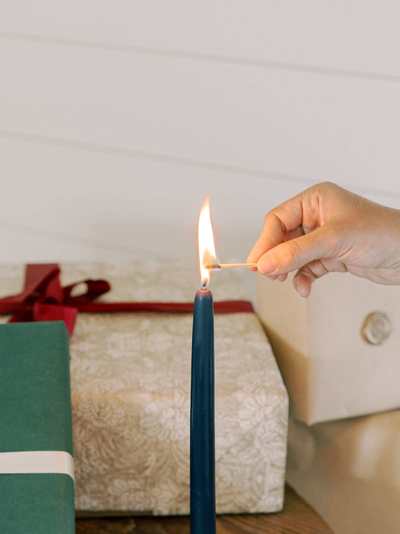 A hand lights a Mo&Co Home - Midnight Blue Dipped Beeswax Taper Candle, surrounded by wrapped gifts, showcasing its handcrafted elegance and eco-friendly design.