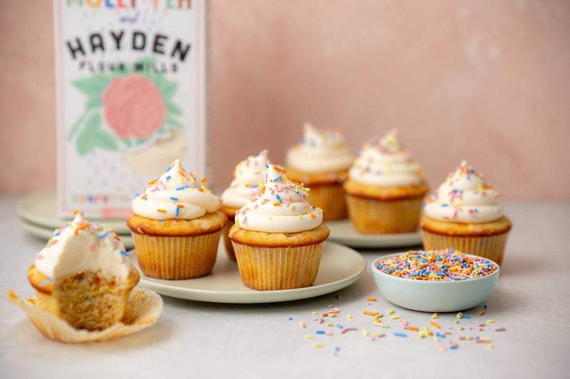 Vanilla cupcakes with buttercream frosting and rainbow sprinkles, featuring Hayden Flour Mills - Molly Yeh x HFM Confetti Cake Mix, highlighting a partially eaten cupcake and sprinkle bowl.