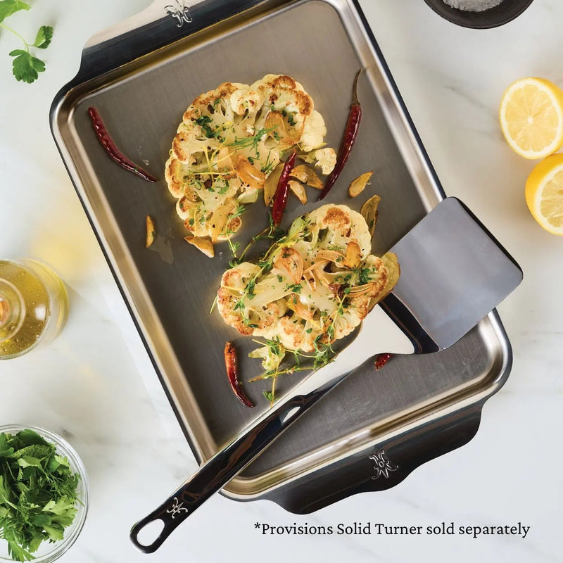Two golden cauliflower steaks with garlic and chilies rest on the Hestan Provisions | Oven Bond Tri-Ply Quarter Sheet Pan, with a metal turner nearby.