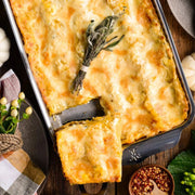 Hestan | Oven Bond Tri-Ply Bakeware, Rectangular Baker holding a cheesy, layered casserole with a portion removed, showing golden crust and herb garnish on rustic table.