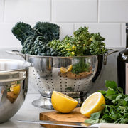 Hestan Provisions Stainless Steel Colander, 5 Quart, filled with leafy greens on a marble countertop, ideal for quick straining and washing vegetables in your kitchen prep.