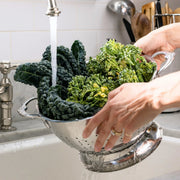 Hestan Provisions | Stainless Steel Colanders, 5 Quart: A person rinses dark leafy greens in a durable, quick-drain colander under a kitchen faucet, ideal for culinary tasks.
