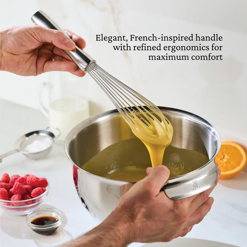 Hestan 12-inch Stainless Steel Whisk mixing batter in a stainless-steel bowl, showcasing its French-style ergonomic handle. Nearby, raspberries, vanilla, powdered sugar, milk, and orange.