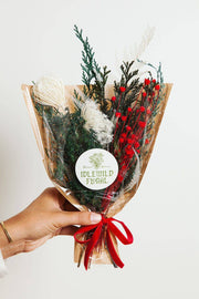 Idlewild Floral Co. - Christmas Mini Market Bouquet, a hand-held dried arrangement with protea, evergreen, and red berries, wrapped in kraft paper and cellophane, tied with a red ribbon.