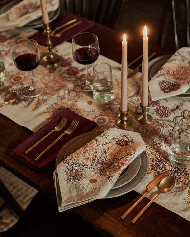 Coral & Tusk - Autumn Blossoms Dinner Napkin elegantly folded on gray-green plates, complements a cozy, fall-inspired table setting with brass accents and lit candles.