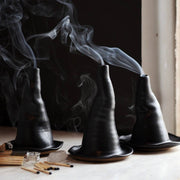 Gravesco Pottery - Witch Hat Incense Burner: Three handcrafted ceramic incense burners, resembling black witch hats, with curling smoke, alongside matches and smudging wood, on a table.