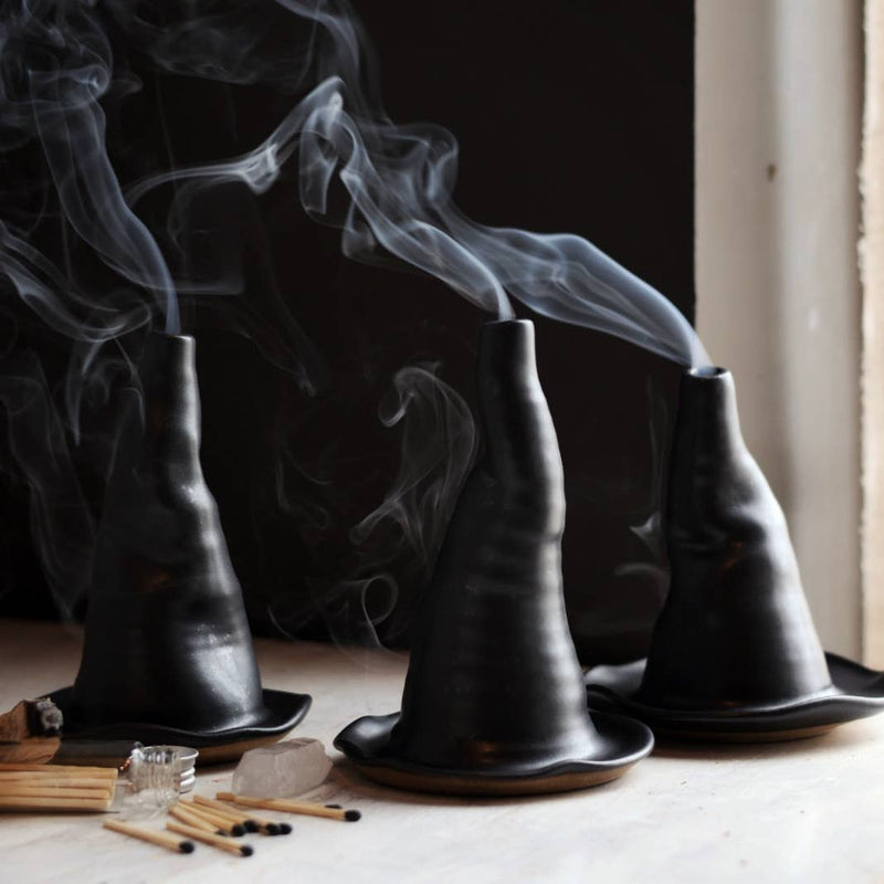 Gravesco Pottery - Witch Hat Incense Burner: Three handcrafted ceramic incense burners, resembling black witch hats, with curling smoke, alongside matches and smudging wood, on a table.