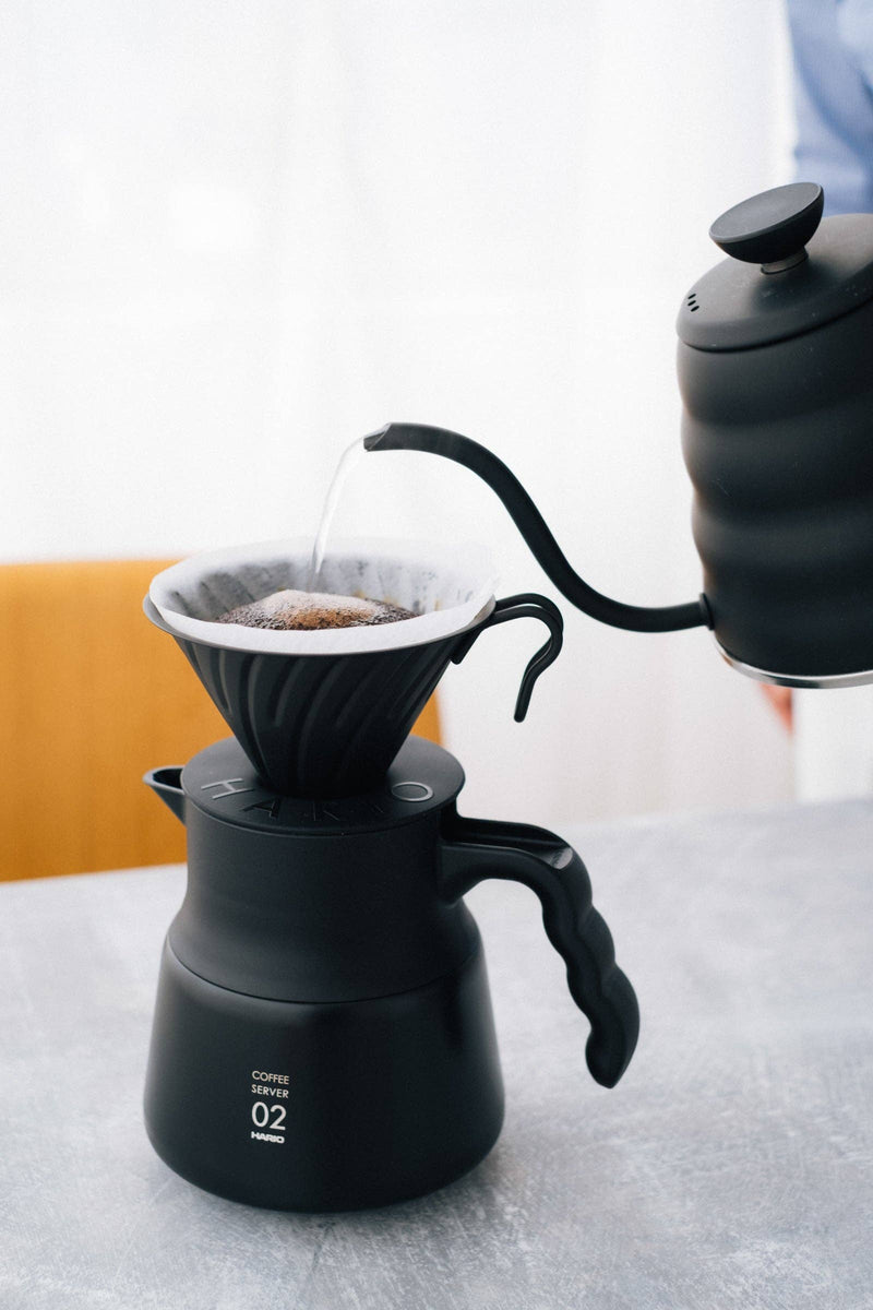 HARIO USA - V60 Metal Dripper: Silver sits atop a matching server. A gooseneck kettle pours water over coffee grounds in a paper filter.