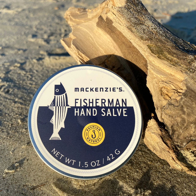 MacKenzie's Fisherman Hand Salve (1.5 oz) tin on sandy ground with driftwood; all-natural, highly moisturizing, perfect for dry skin, made in New England.