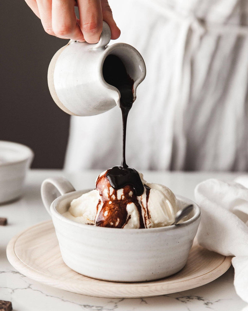 Hand pours warm chocolate sauce from the Farmhouse Pottery - Bell Pitcher over vanilla ice cream, illustrating its elegant design and versatile functionality.