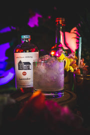 Pink House Alchemy - Lavender Simple Syrup bottle with a house illustration, next to a glass of iced lavender drink with lemon.