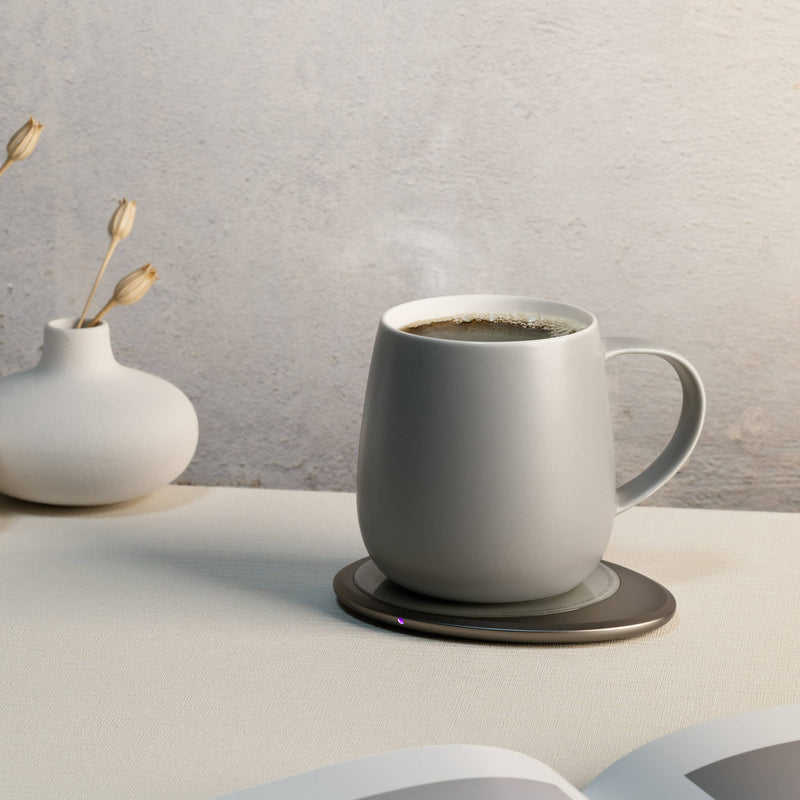 OHOM - Ui 3 Self-heating Mug Set: Jasmine White with steaming coffee on a warming coaster, alongside a white vase with dried buds and an open book corner.