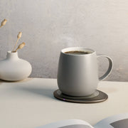 OHOM - Ui 3 Self-heating Mug Set in soft gray, on a warming plate, with a sculptural vase and open book, enhancing your coffee experience.