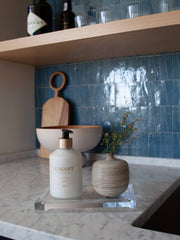 L'AVANT Collective - Fresh Linen Hand Lotion in an elegant glass dispenser, displayed alongside a vase with flowers on a counter.