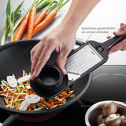 A person uses the GEFU | Violi 2.0 Adjustable Mandoline to grate vegetables into a pan, highlighting its functionality for kitchen tasks.