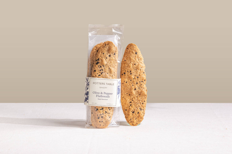 Potters Table - Cured Olive & Cracked Pepper Flatbreads, one packaged, one displayed, showcasing their hand-stretched, speckled texture, ideal for charcuterie or cheese spreads.
