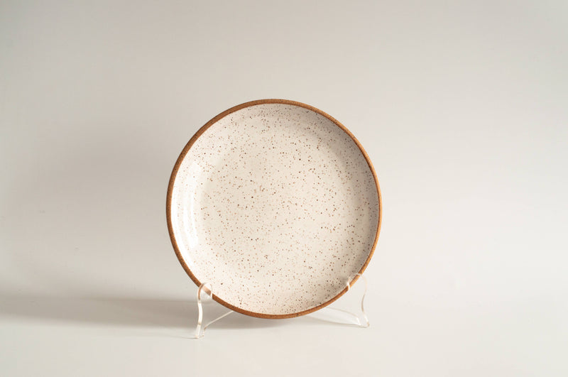 RachaelPots - Salad Plate: Nude/White, a speckled ceramic plate with an earthy rim, handcrafted in Brooklyn, displayed upright on a clear stand.