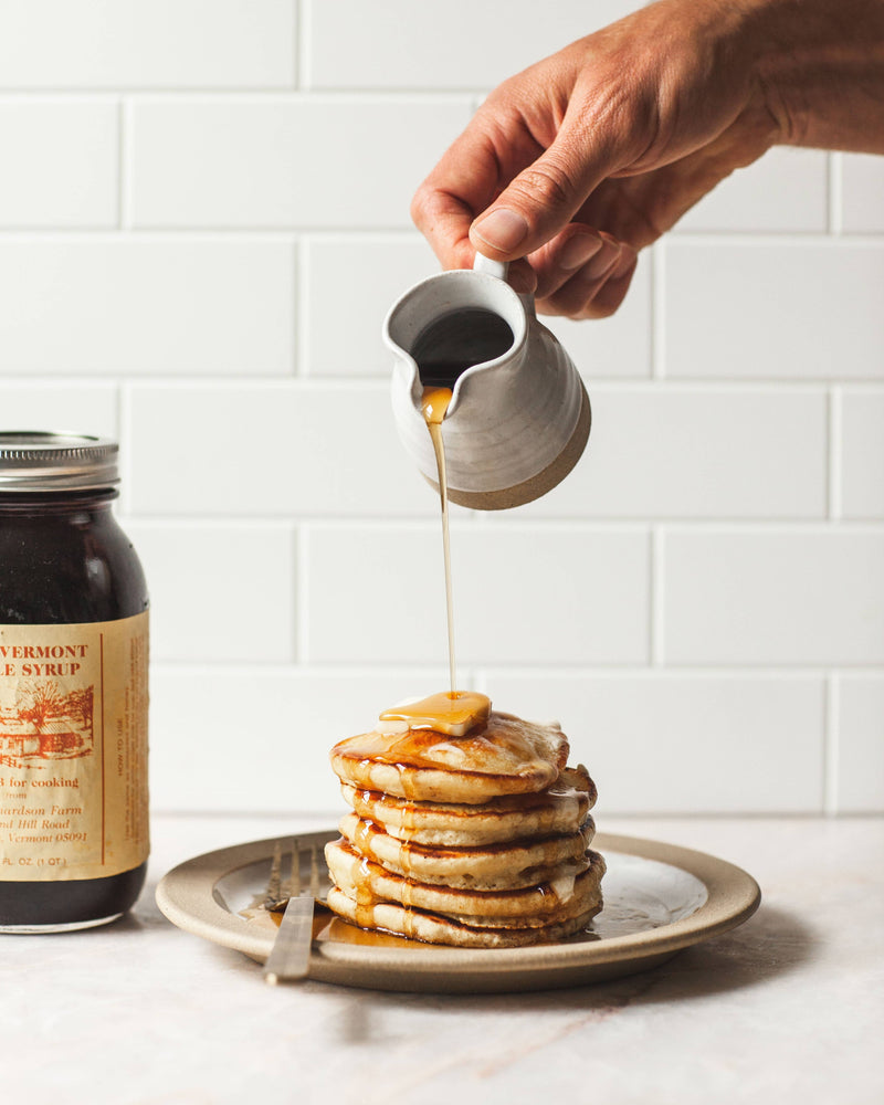 Farmhouse Pottery Bell Pitcher elegantly drizzles Vermont Maple Syrup over pancakes, showcasing its charm and functionality for serving dressings or cream.