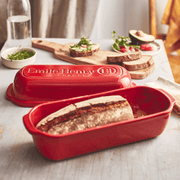 Emile Henry Loaf Bread Baker with freshly baked crusty bread on a wooden tabletop, surrounded by sliced bread, avocado toasts, and kitchen essentials.