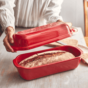 A person lifts the lid off an Emile Henry Loaf Bread Baker, revealing a freshly baked artisan loaf inside the burgundy ceramic cloche.