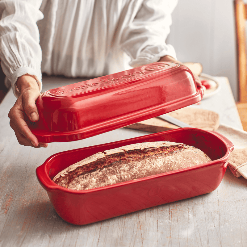 A person lifts the lid off an Emile Henry Loaf Bread Baker, revealing a freshly baked artisan loaf inside the burgundy ceramic cloche.
