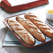 Emile Henry | Baguette Baker, Burgundy: Three freshly baked baguettes in a red three-slot pan, showcasing crusty texture; a butter dish in the background.