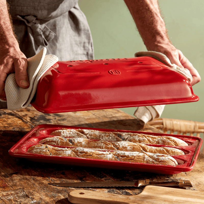 Hands lift the lid of an Emile Henry | Baguette Baker, Burgundy, revealing three freshly baked baguettes on a rustic table with a bread knife and cutting board.