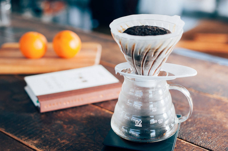HARIO USA - HARIO V60 Glass Dripper 02: Olive Wood displayed in a pour-over setup on a wooden table with a brewing carafe and scale, enhancing coffee preparation.