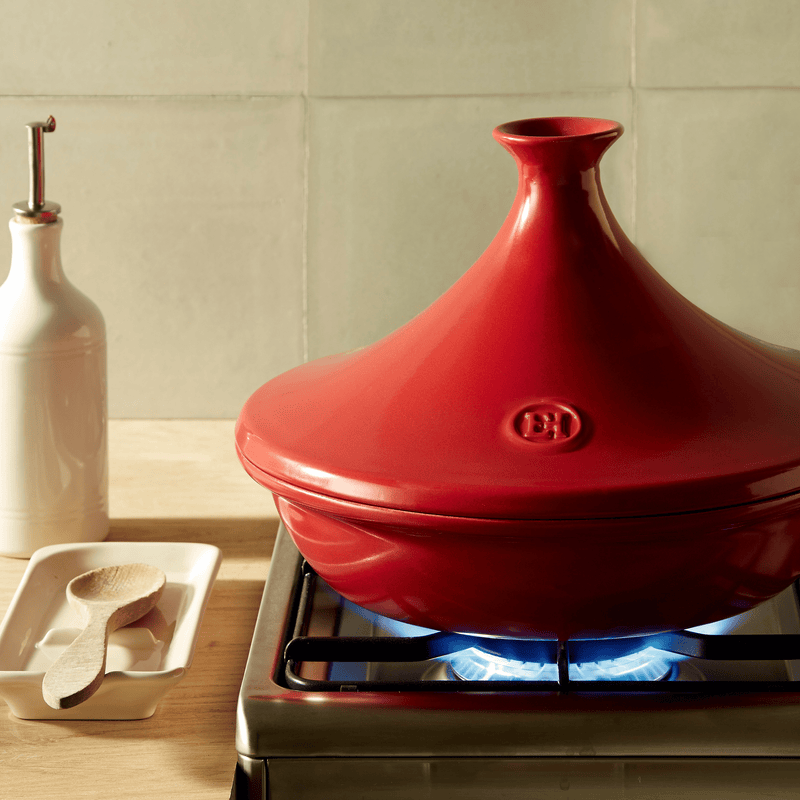 Emile Henry Tagine, Charcoal, sits on a lit stove, showcasing its unique design for simmering stews, surrounded by a white oil cruet and wooden spoon.