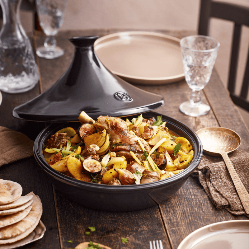 Emile Henry | Tagine, Charcoal reveals chicken stew with vegetables and figs, surrounded by flatbreads and dining essentials on a rustic table, showcasing culinary craftsmanship.
