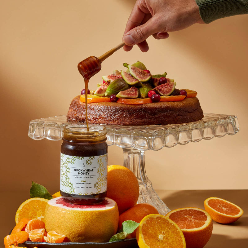 Villa Jerada - Buckwheat Honey - 1lb drizzled over a fig and citrus-topped cake on a stand, surrounded by citrus fruits and a labeled jar on a grapefruit.