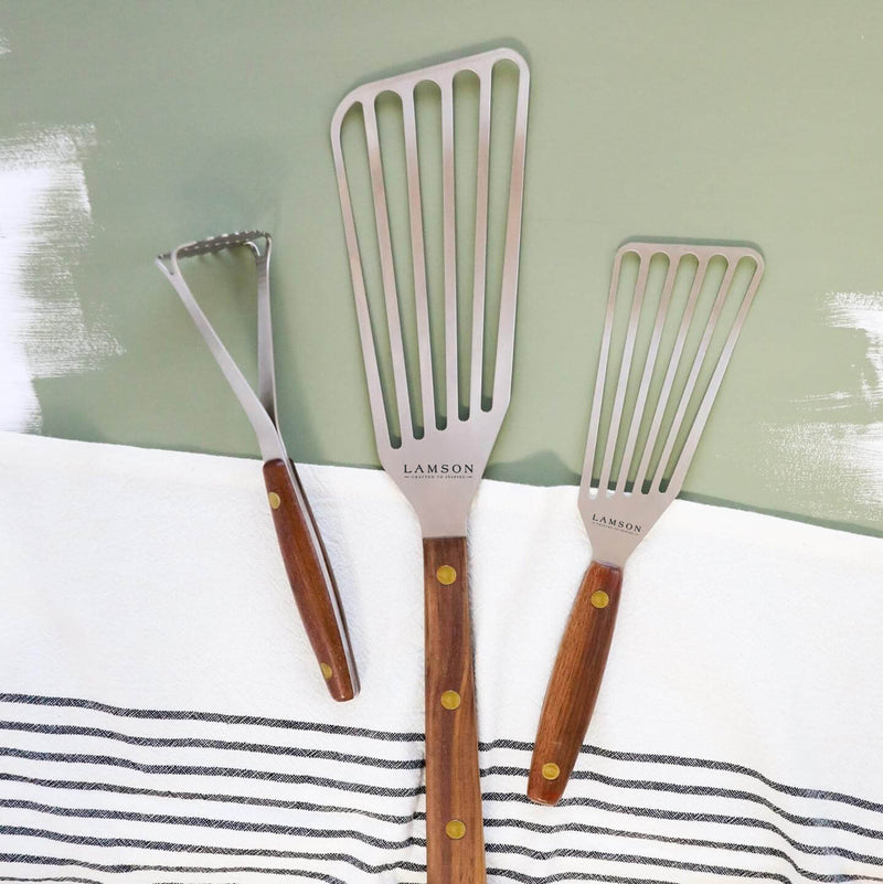 Lamson | Vintage Mini Avocado + Spud Masher surrounded by spatulas and tongs on a towel, showcasing traditional kitchenware for effective mashing tasks.