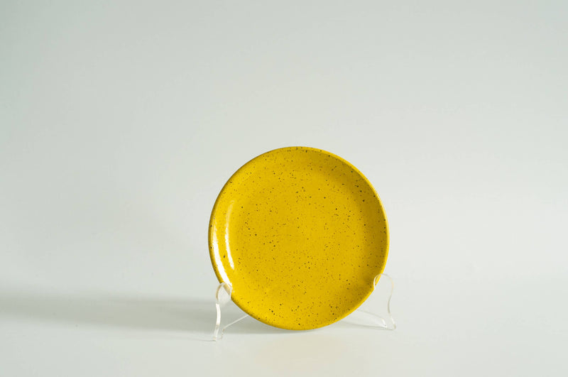 RachaelPots - Dessert Plate: Sage, a mustard-yellow ceramic plate with black speckles, displayed on a stand, ideal for desserts. Handcrafted stoneware, microwave and dishwasher safe.