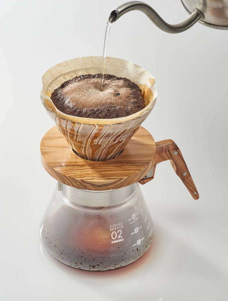 HARIO USA - HARIO V60 Glass Dripper 02: Olive Wood, showcasing a pour-over coffee brewing with a gooseneck kettle, highlighting its heat-resistant glass and wooden collar.